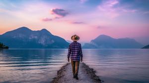 Man with purple checkered shirt short hair wearing a brimmed explorer's hat walking across lakeside in northern Italy Lake garda at sunrise.