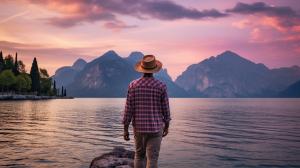 Man with purple checkered shirt short hair wearing a brimmed explorer's hat walking across lakeside in northern Italy Lake garda at sunrise.