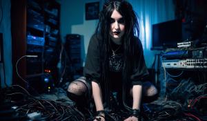 Mature goth girl kneeling on bed in appartment, computers cables and wires scattered around the room aswell as bulky desktop computers with code on them, she is front facing veiwer,close up shot, ,wearing grunge 90s clothing, pallid skirt, soft grunge makeup,