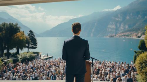 Man standing in a suit with rostrum and stand with microphone on it, many Jehovah's witnesses gathered for exercise patience 2023 convention of Jehovah's Witnesses at a outdoor venue at Lake Garda Northern Italy.