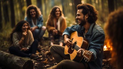 Young people having fun around a campfire. Drinking, playing guitar and laughing a lot. They are in a clearing in a wood, with tents around them.